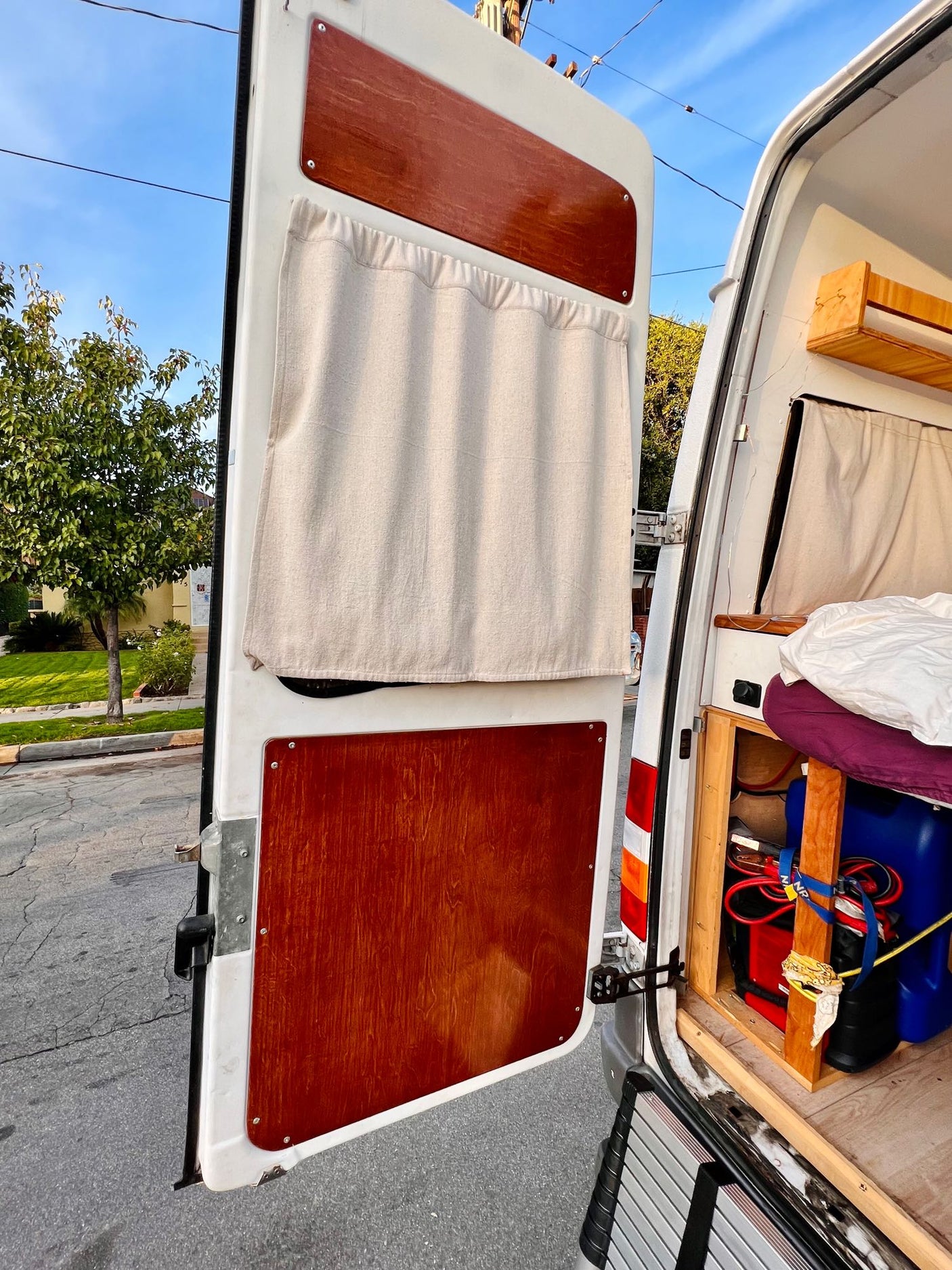 Open rear doors of an rv rental camper van reveal a custom interior: wood panels, fabric curtains, and a cozy bed.