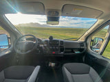 Inside an RV rental: dashboard, seats, and a vast sunny view of mountains, green fields, and sky. Road trip ready.