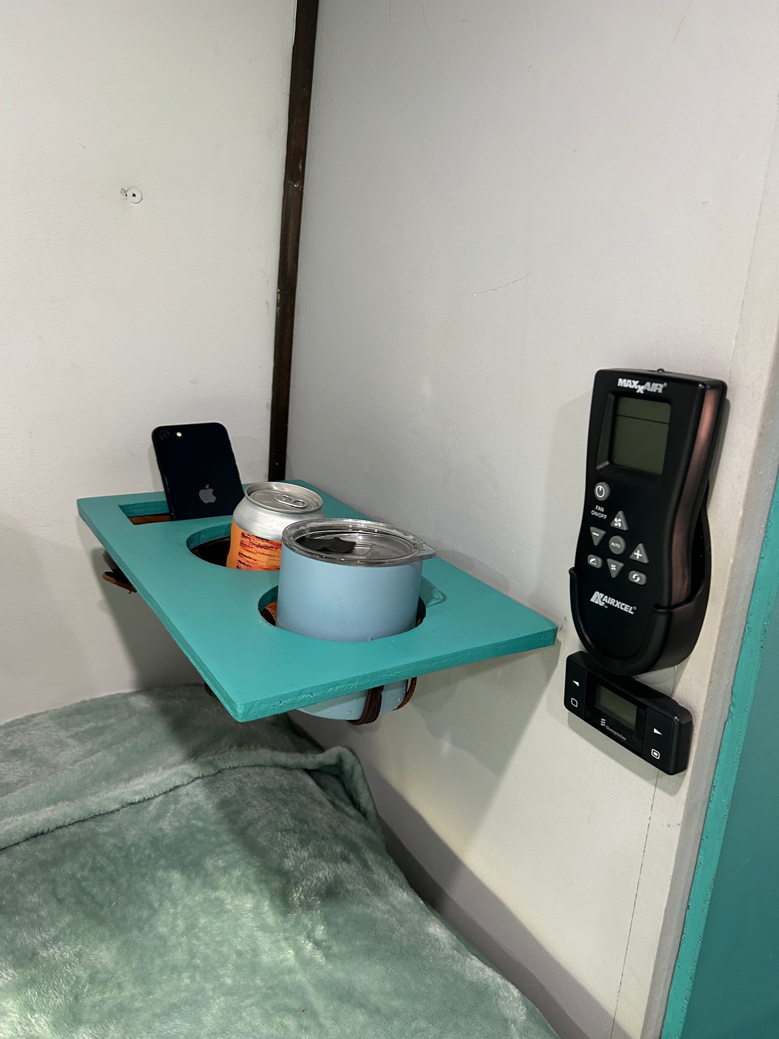 Cozy RV rental bedroom with a teal bedside shelf holding a phone, canned drink, and mug, alongside climate controls.