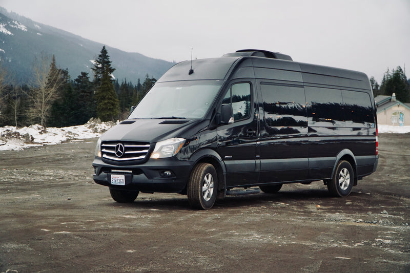 A sleek black Mercedes-Benz Sprinter van, likely an RV rental, parked on a snowy dirt road with mountains and trees. — photo 2