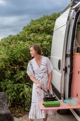 Enjoying an RV rental: woman cooking outdoors with a portable stove next to a white campervan, surrounded by green foliage.