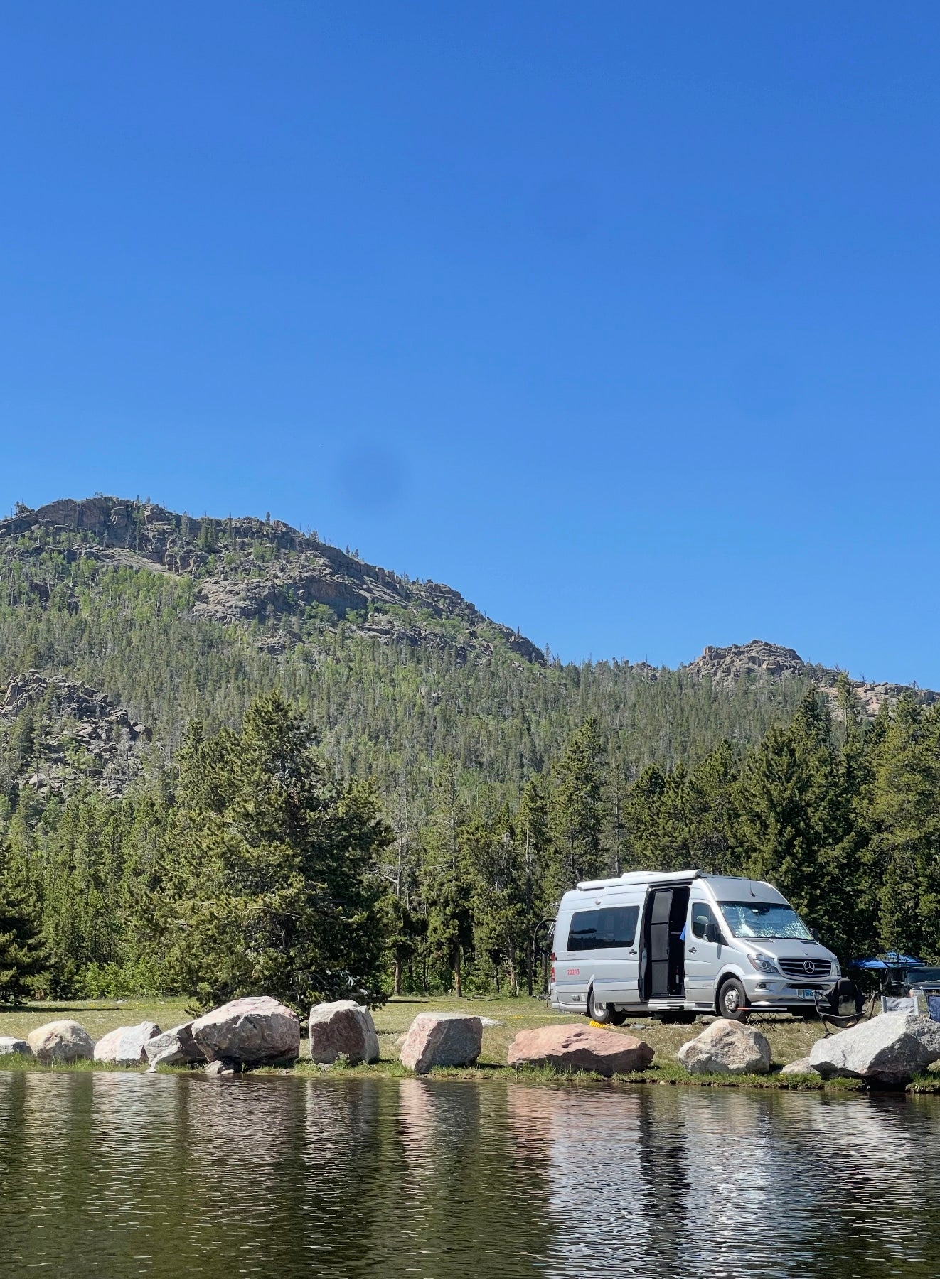 Scenic RV rental adventure: a silver camper van parked beside a calm lake, backed by forested mountains under a blue sky.