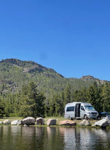 Scenic RV rental adventure: a silver camper van parked beside a calm lake, backed by forested mountains under a blue sky.