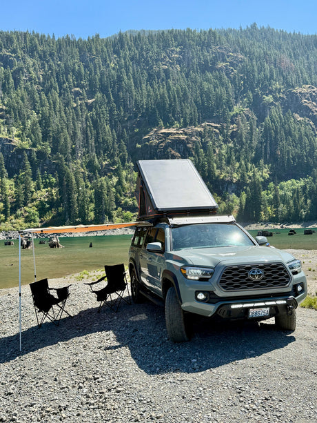 A grey Toyota Tacoma with rooftop tent & side awning parked on a gravel shore by a lake with a forest background. Ideal for an RV rental adventure.