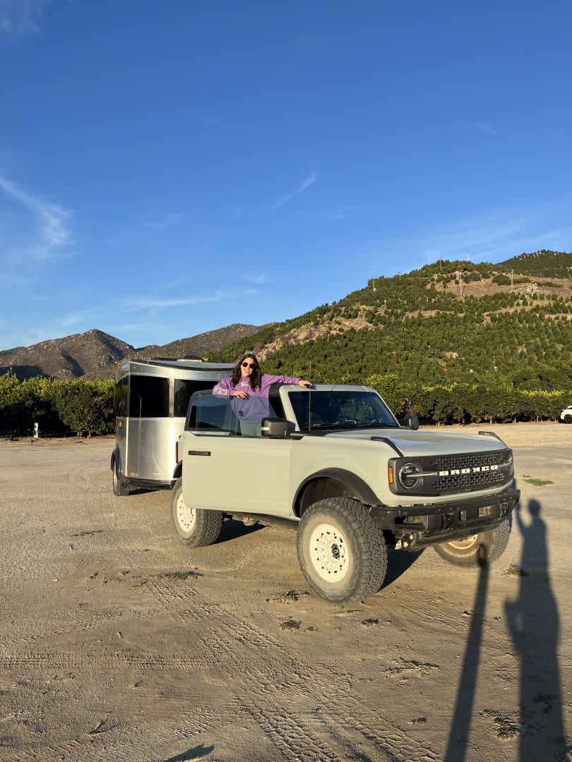 Woman in a light Ford Bronco SUV, doors removed, towing a silver RV rental for a mountain adventure.