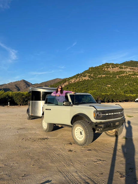 Woman in a light Ford Bronco SUV, doors removed, towing a silver RV rental for a mountain adventure.