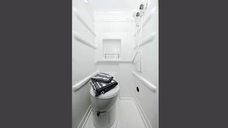 Compact white RV rental bathroom with toilet, wall-mounted shower, and shelf. Towels are neatly folded on the toilet.