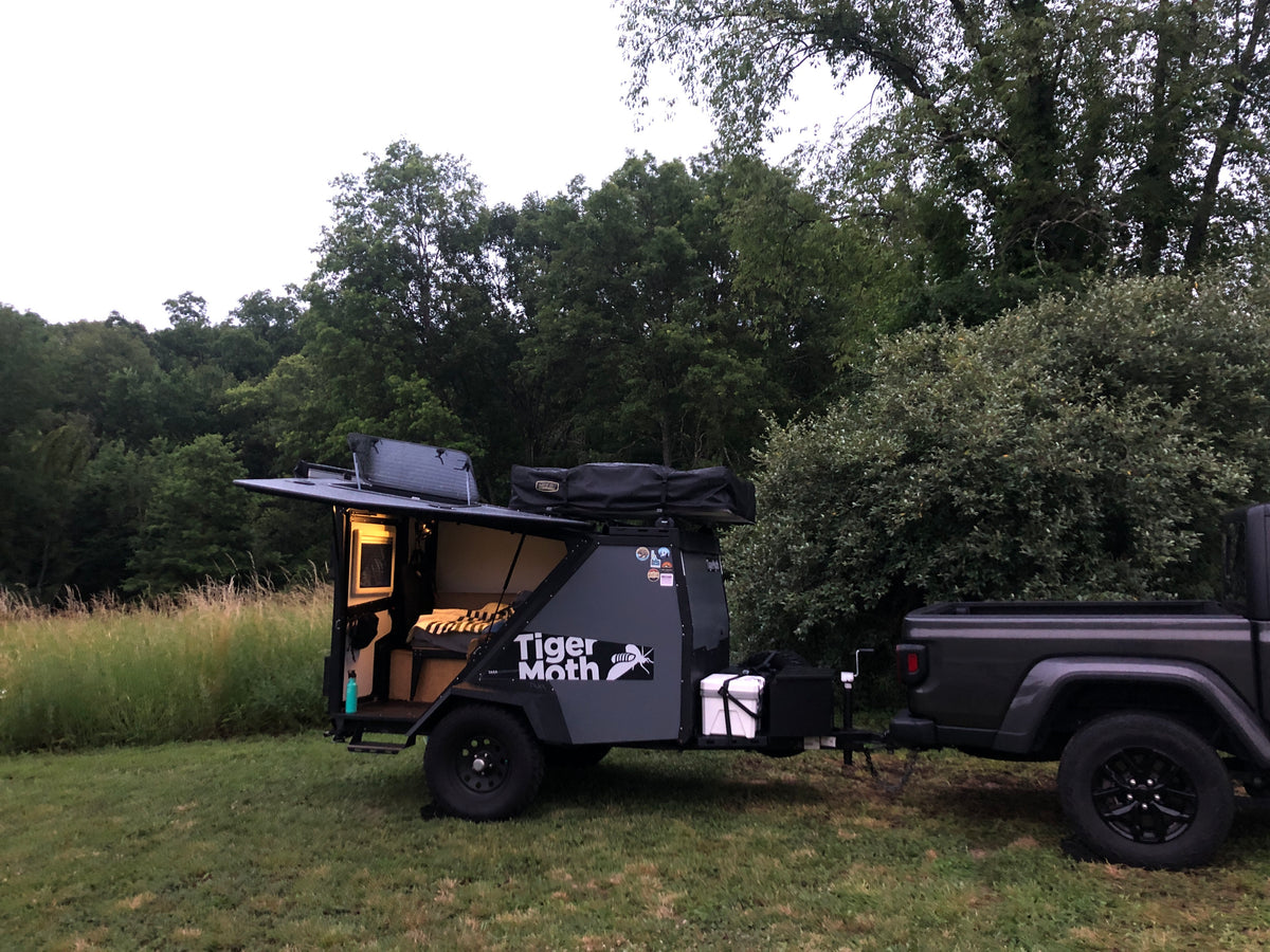 This compact 'Tiger Moth' camping trailer rv rental, pulled by a pickup truck, is set up in a lush, green outdoor spot.