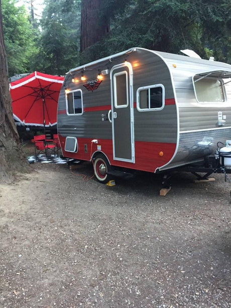 This retro RV rental, a gray and red travel trailer, is perfectly set up for outdoor camping with a red umbrella and chairs.