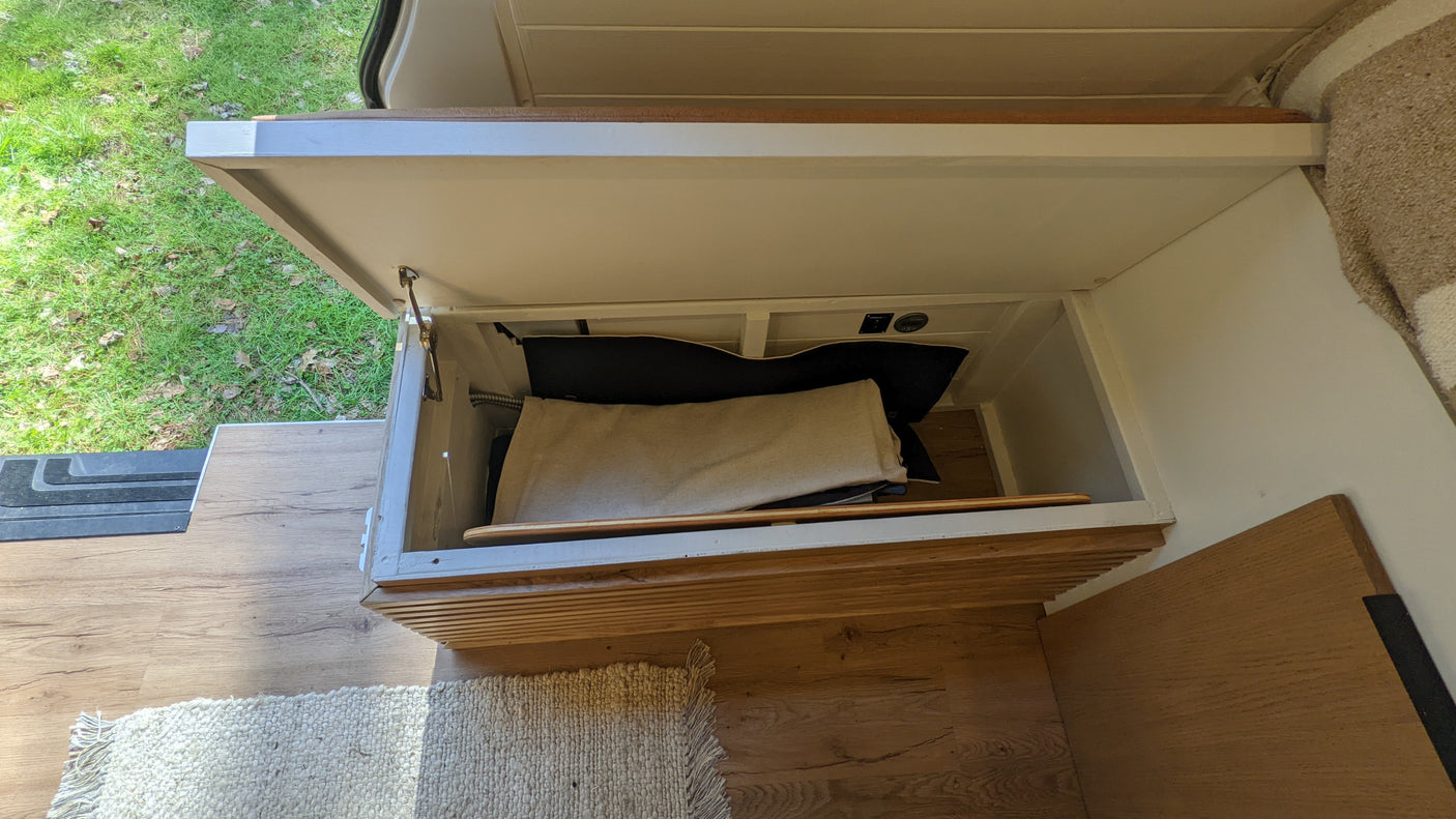 RV rental: Open storage bench with a beige blanket, black bag, and electrical outlets visible inside a camper van interior.
