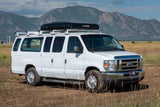 White Ford RV rental van with a hardshell rooftop tent, parked in a scenic field with mountains. Ideal for adventure travel.