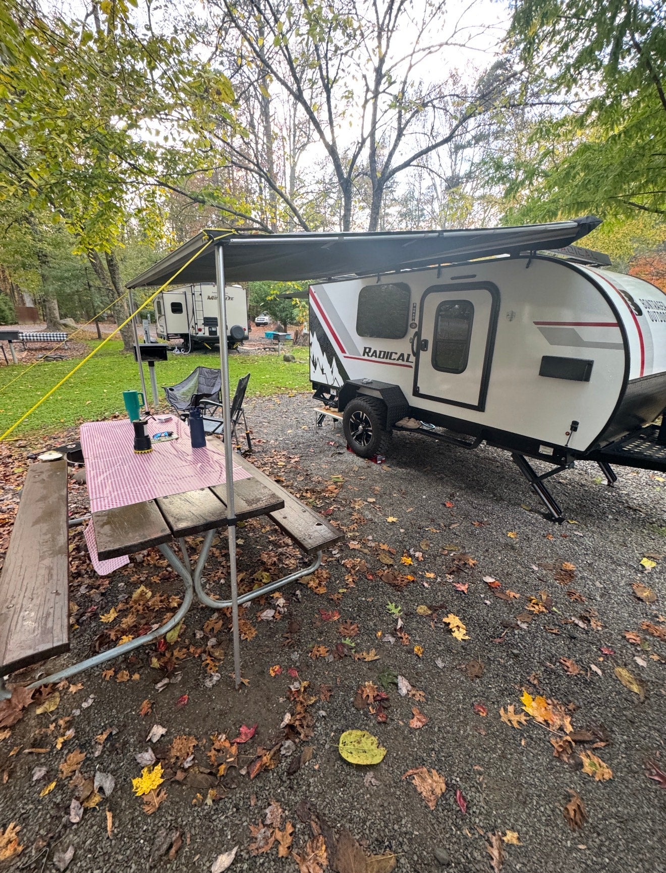 RV rental teardrop camper with awning & picnic table at a fall campsite among colorful leaves.