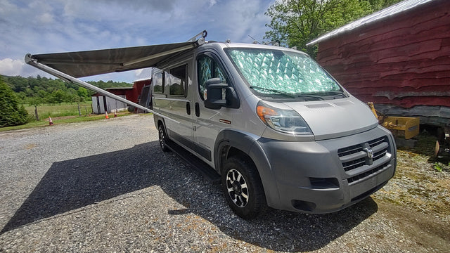 Explore with this silver Ram ProMaster rv rental! Awning extended, parked on gravel with a sunshade, ready for scenic trips.
