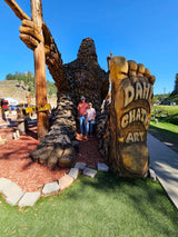 A couple poses with a huge Bigfoot wood carving, featuring Dahl Chat Art. Ideal stop for your RV rental journey!