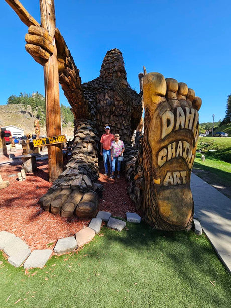 A couple poses with a huge Bigfoot wood carving, featuring Dahl Chat Art. Ideal stop for your RV rental journey!