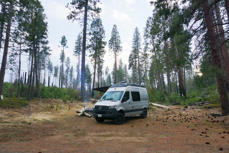 Enjoy nature's beauty with an RV rental. Silver camper van parked in a forest clearing next to a smoky campfire.
