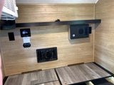 Inside an RV rental: wall-mounted AC, digital control panel, electrical components, and hinged bed base with wood-look flooring.