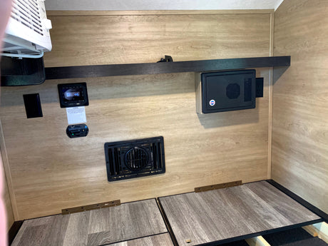 Inside an RV rental: wall-mounted AC, digital control panel, electrical components, and hinged bed base with wood-look flooring.