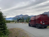 A red rv rental van faces distant Alaskan mountains, a glacier lake, and a rest stop building under a cloudy sky.