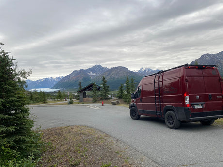 A red rv rental van faces distant Alaskan mountains, a glacier lake, and a rest stop building under a cloudy sky.