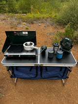 RV rental ready: Portable camping kitchen setup with a Coleman stove, pot, griddle, fan, and mugs on a folding table.