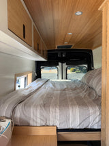 RV rental bedroom interior: cozy bed with striped linens, bamboo ceiling & cabinets, white walls, and rear windows.