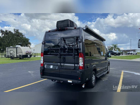 Black Tellaro camper van with bike rack and awning, perfect for RV rental, parked at Lazydays RV. A travel trailer is in the background.