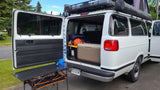White camper van with open rear doors showing gear, cooler, and a table. Ideal for an RV rental adventure.