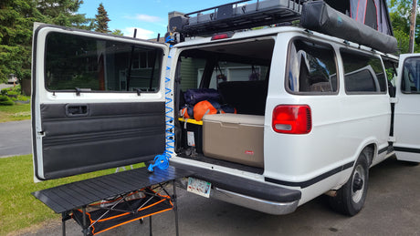 White camper van with open rear doors showing gear, cooler, and a table. Ideal for an RV rental adventure.