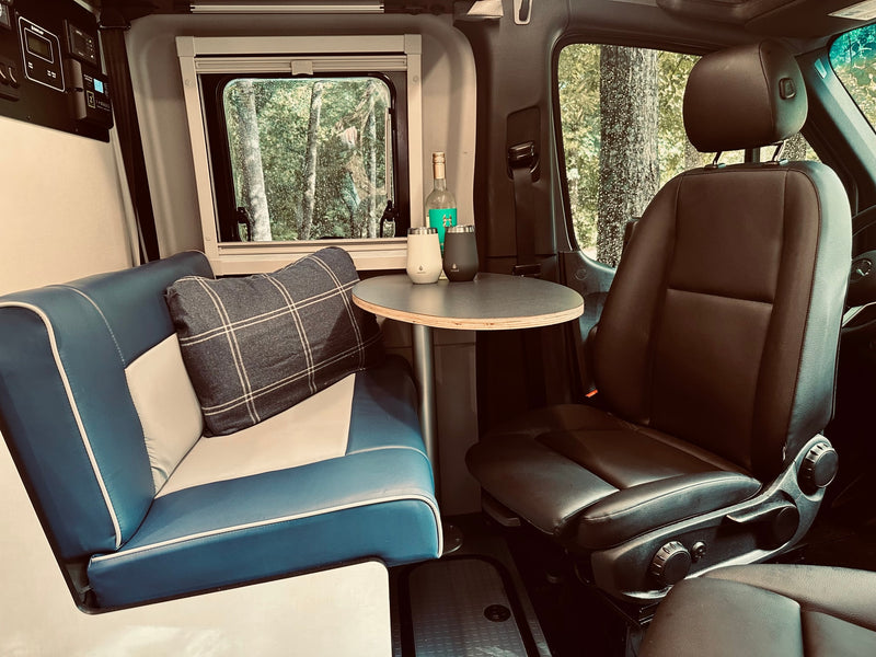 Step inside this comfortable RV rental. Features a blue bench seat, round table with drinks, and a serene forest view. — photo 4