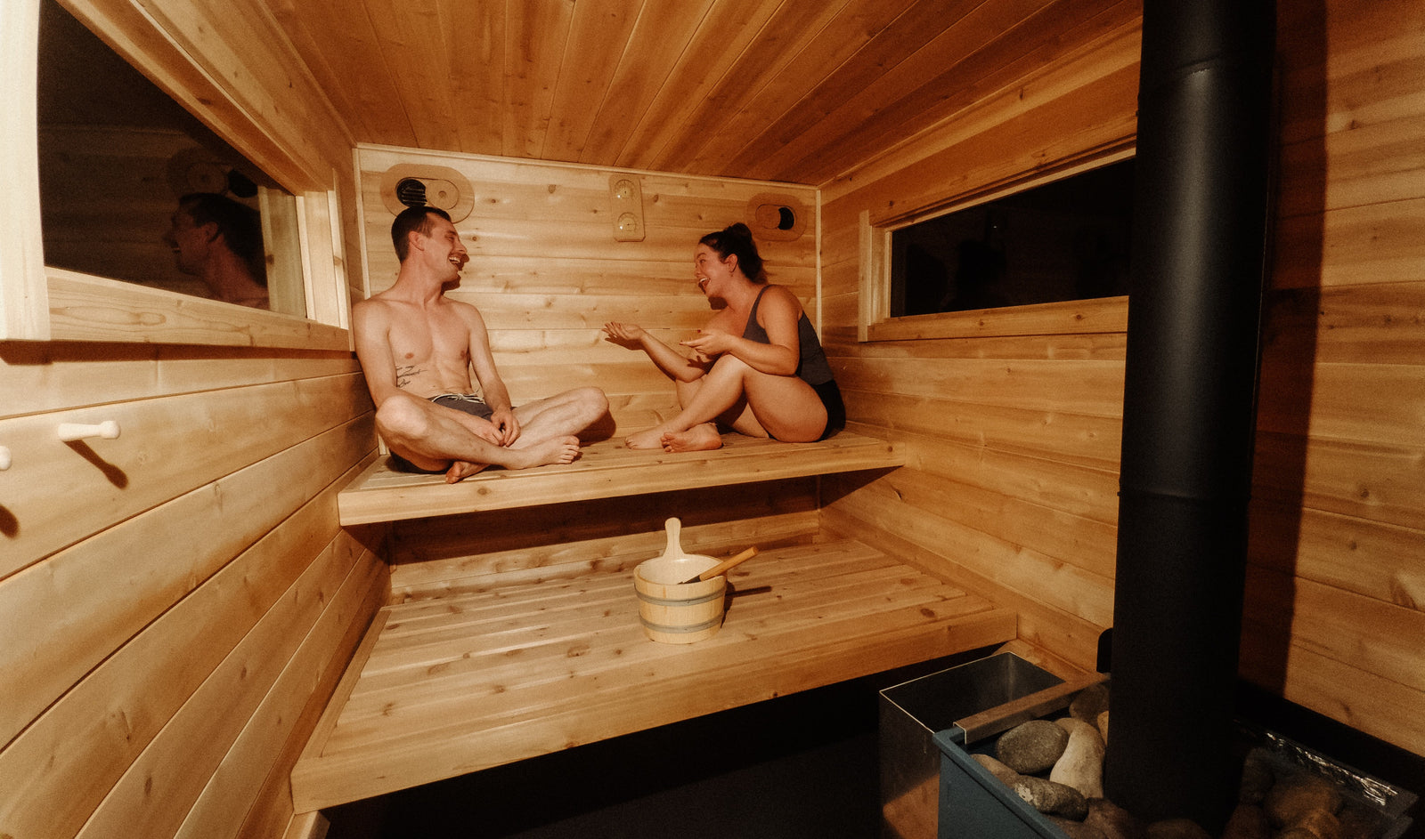 RV rental relaxation: Two people chat and laugh in a cozy wooden sauna with a heater and bucket, perfect for unwinding.
