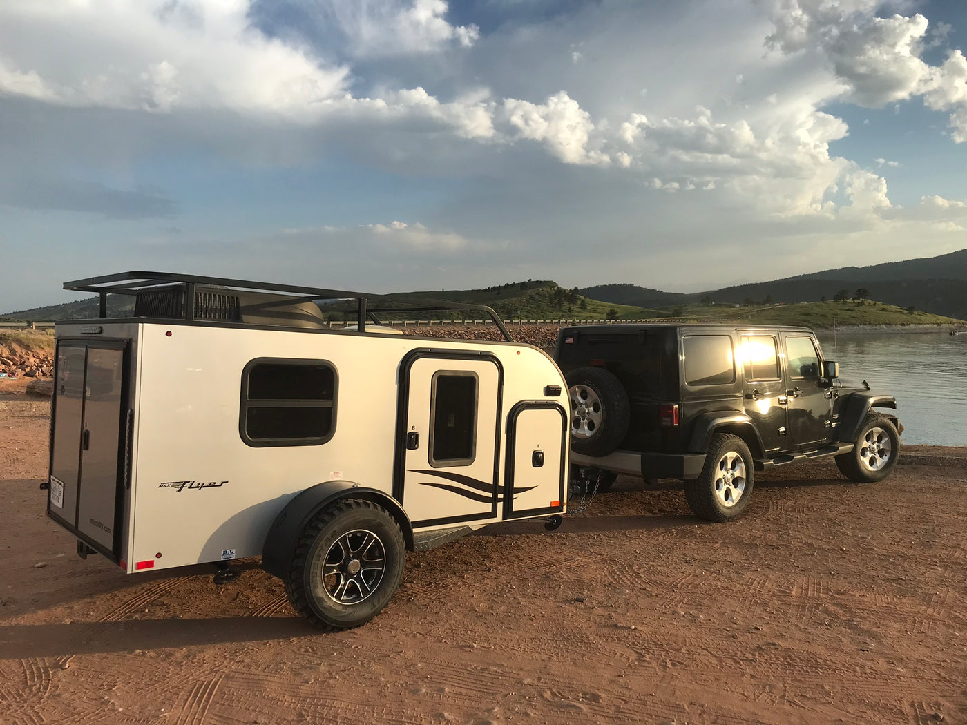 Black Jeep Wrangler towing a white inTech Max Flyer RV, parked lakeside. An ideal RV rental for off-road camping adventures.