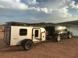 Black Jeep Wrangler towing a white inTech Max Flyer RV, parked lakeside. An ideal RV rental for off-road camping adventures.