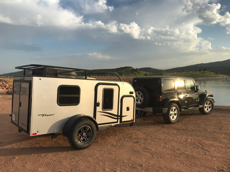 Black Jeep Wrangler towing a white inTech Max Flyer RV, parked lakeside. An ideal RV rental for off-road camping adventures.