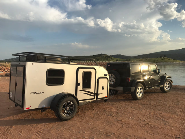 Black Jeep Wrangler towing a white inTech Max Flyer RV, parked lakeside. An ideal RV rental for off-road camping adventures.