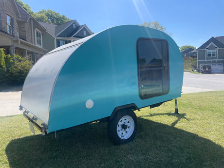 Sunny day shows a compact, light blue and silver teardrop camper, ideal for an RV rental, parked on green grass.