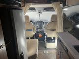 Inside an RV rental, looking forward past a refrigerator and TV towards the front seats and cockpit dashboard.