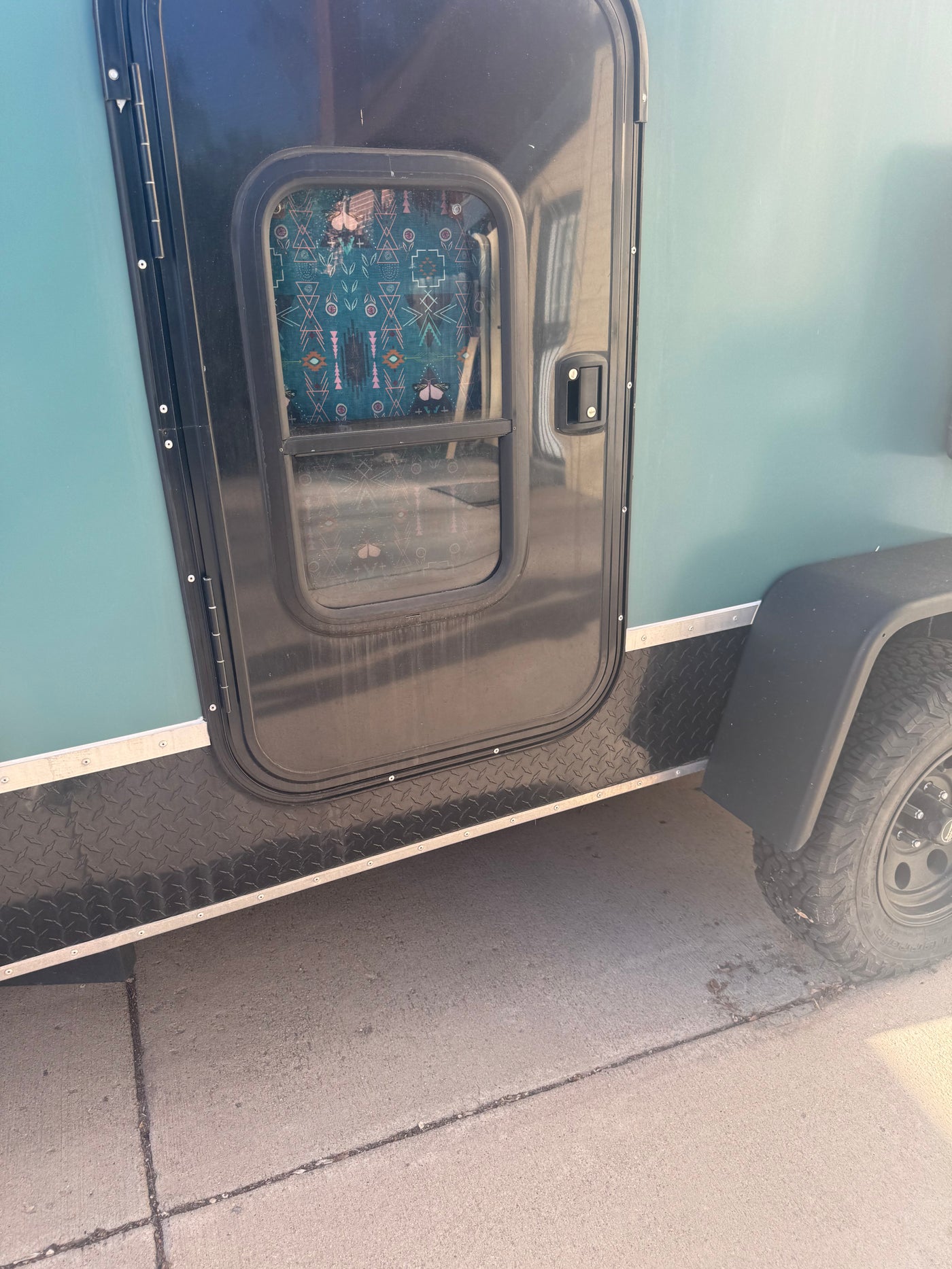 Close-up of a teal RV rental's side, showing its black door, diamond plate trim, and rugged all-terrain tire. — photo 11