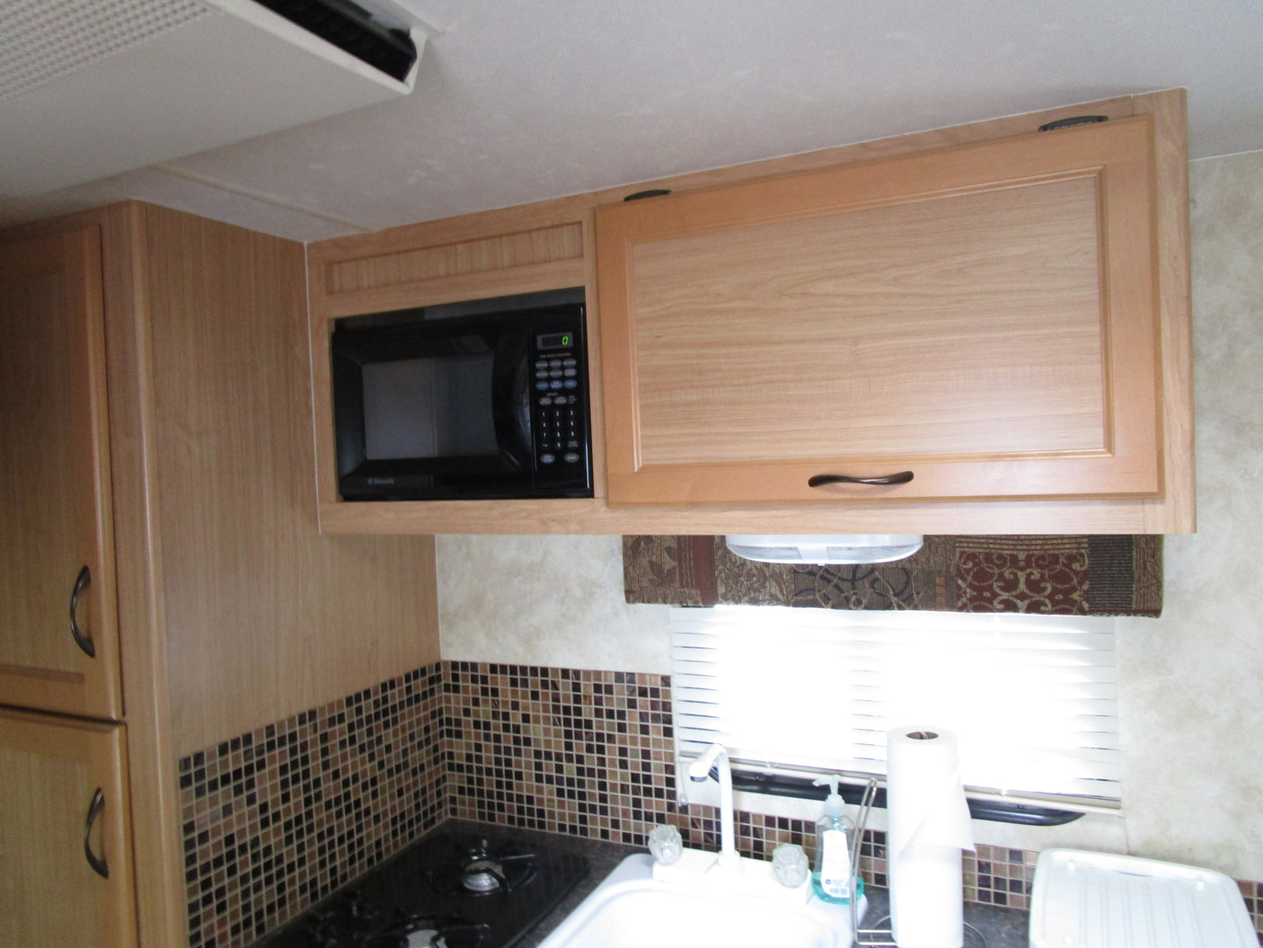 Experience comfort in this RV rental kitchen, featuring wood cabinets, microwave, tiled backsplash, stovetop, and sink.