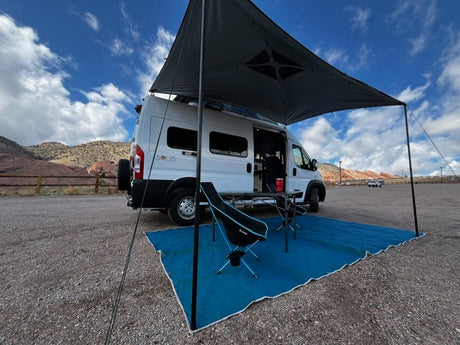White RV rental camper van with extended awning, chairs, and mat on a gravel lot. Scenic mountain backdrop.
