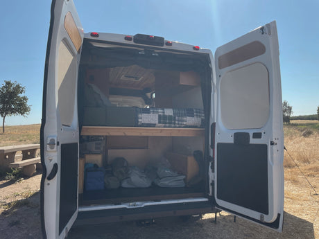 Open rear doors of a white camper van showing its interior bed & storage, ready for adventure. Ideal for an RV rental.