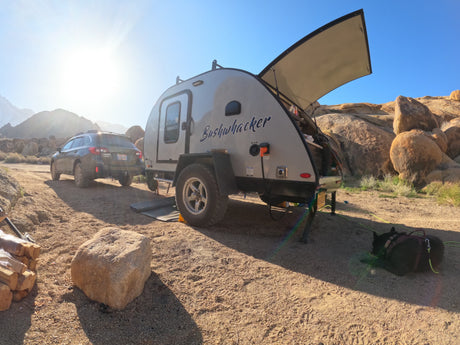 Subaru towing a Bushwhacker teardrop RV rental, set up for camping in a sunny, rocky desert landscape with a resting dog.