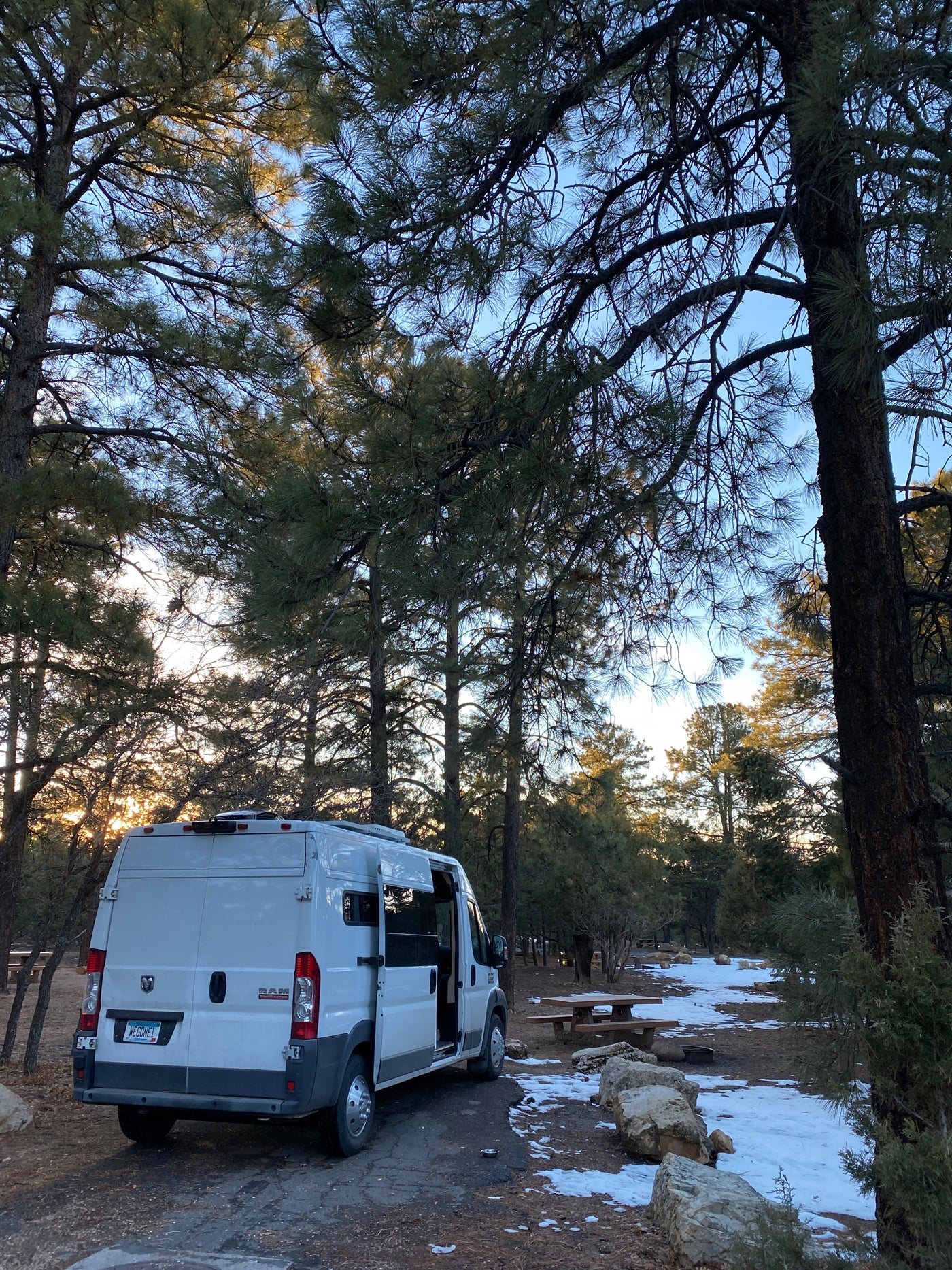 A white Ram ProMaster camper van, likely an rv rental, is parked at a tranquil snowy campsite under tall pine trees at dusk. — photo 10