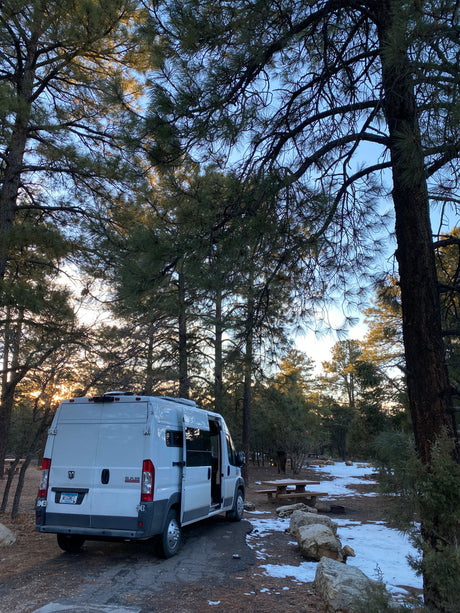 A white Ram ProMaster camper van, likely an rv rental, is parked at a tranquil snowy campsite under tall pine trees at dusk.