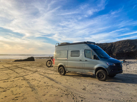 Experience van life! This Winnebago Revel RV, perfect for an RV rental, is parked on a golden sandy beach at sunset with a bike.
