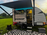 An RV rental travel trailer is set up with an illuminated awning, outdoor mat, grill, and cooler for a relaxing evening.