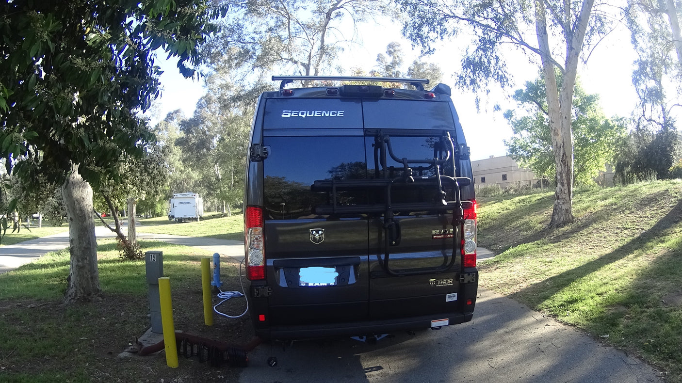 Rear view of an RV rental, a black campervan with a bike rack, parked at a sunny campsite 15, ready for travel.