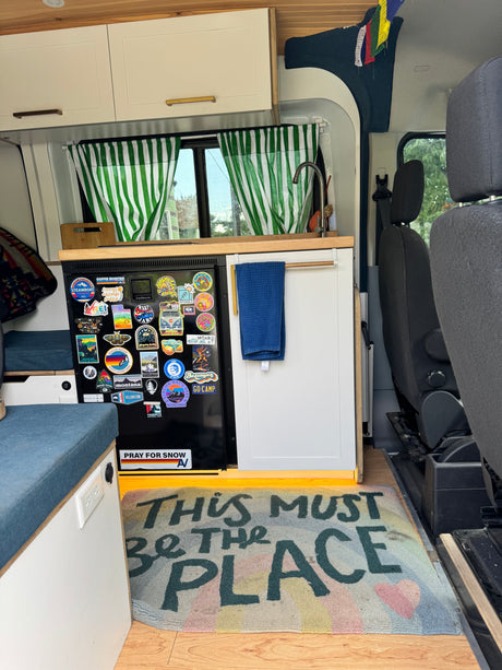 Cozy RV rental interior featuring a kitchen with travel stickers, a "THIS MUST BE THE PLACE" rug, and custom cabinetry.