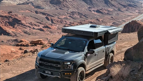 A grey Ford Raptor pickup with an RV rental truck camper navigates a dirt trail in a red rock desert landscape.