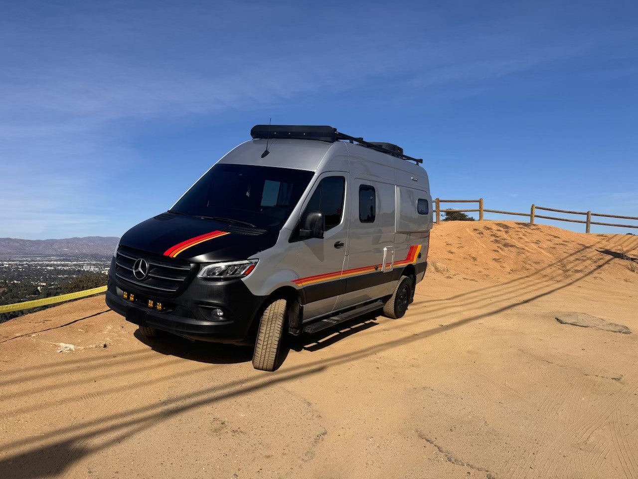 This sleek RV rental, a custom gray Mercedes Sprinter camper van with bold stripes, is ready for adventure on a dirt hilltop.